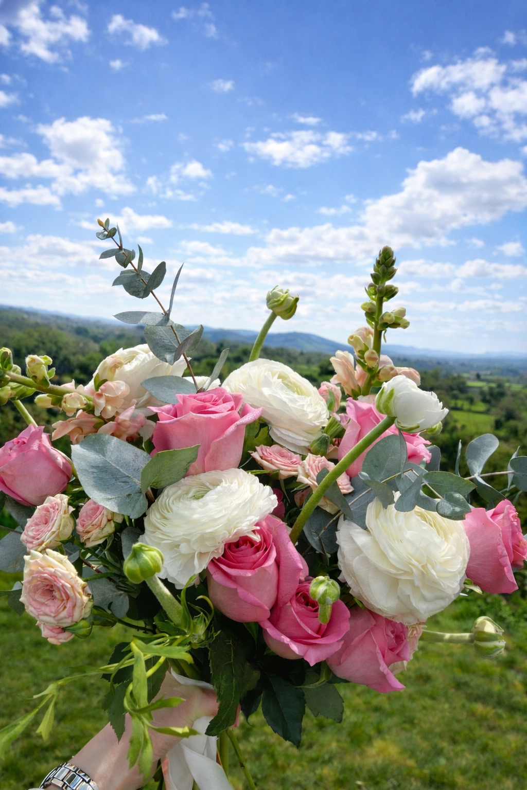 A vibrant bouquet of pink and white roses, with some eucalyptus and other green foliage, held up against a scenic countryside landscape with a blue sky and scattered white clouds.