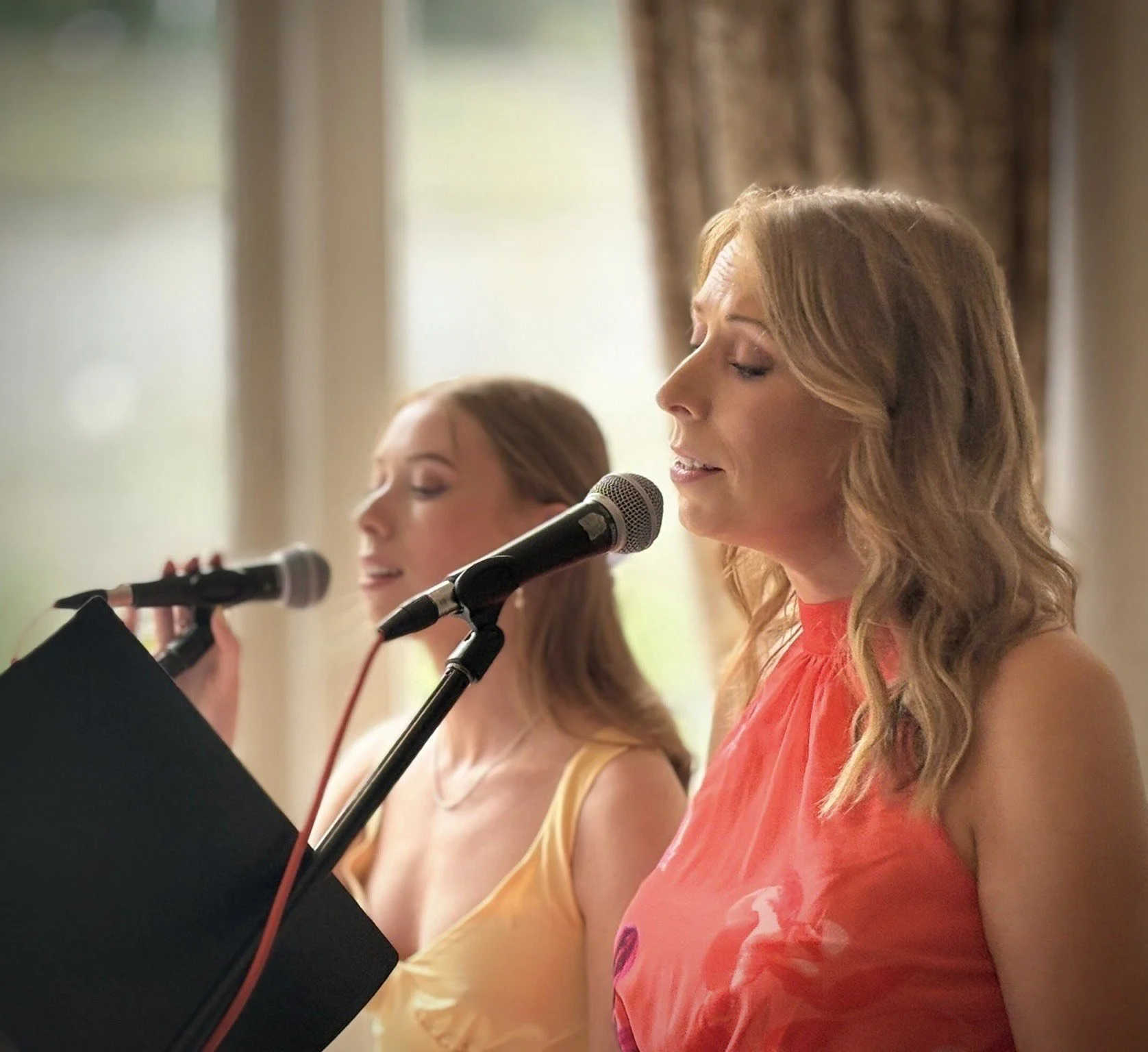 Two women singing into microphones during a performance in a cozy indoor setting with curtains and windows in the background.
