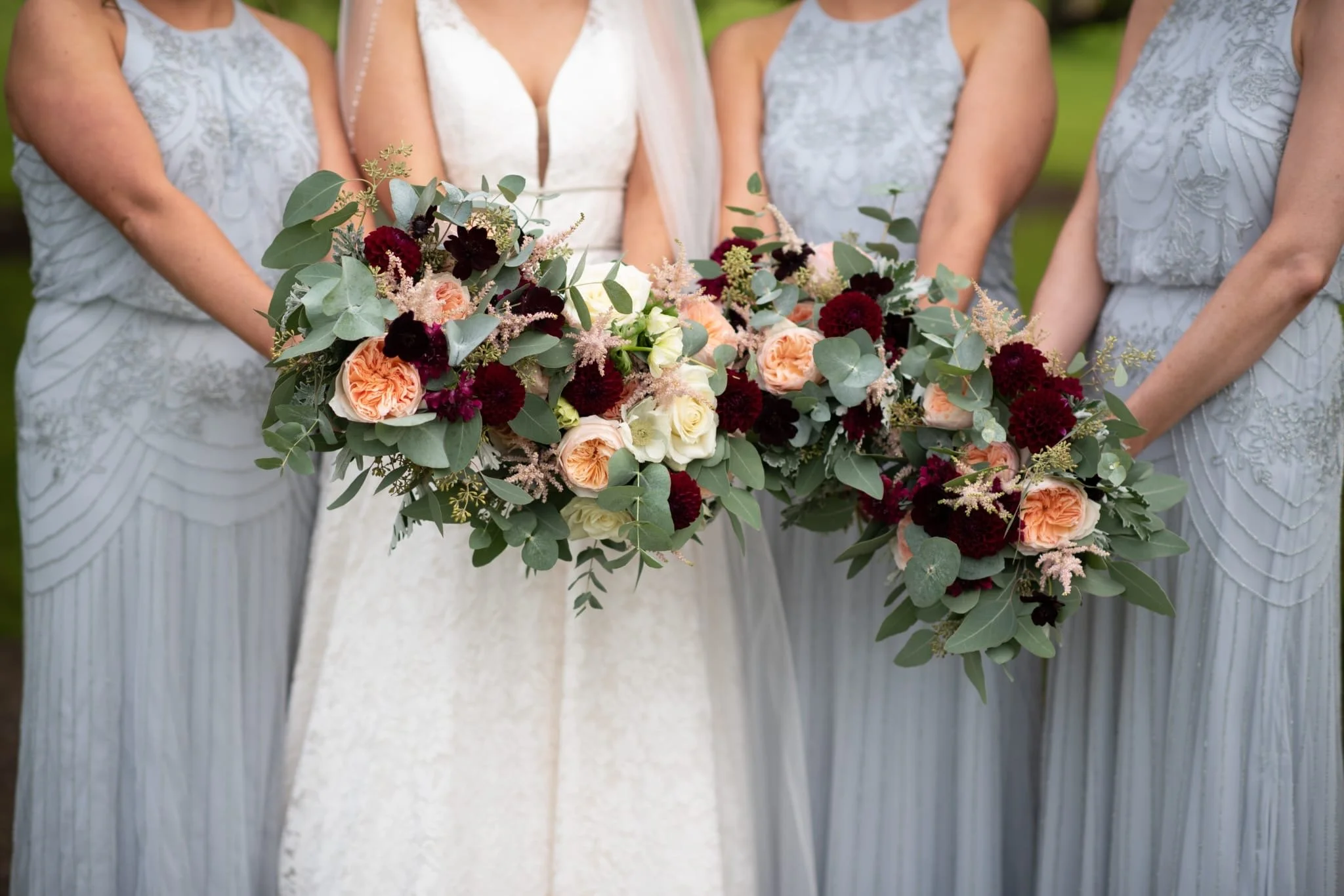A bride and her bridesmaids holding a large, cascading bouquet of peach, cream, and burgundy flowers with greenery.