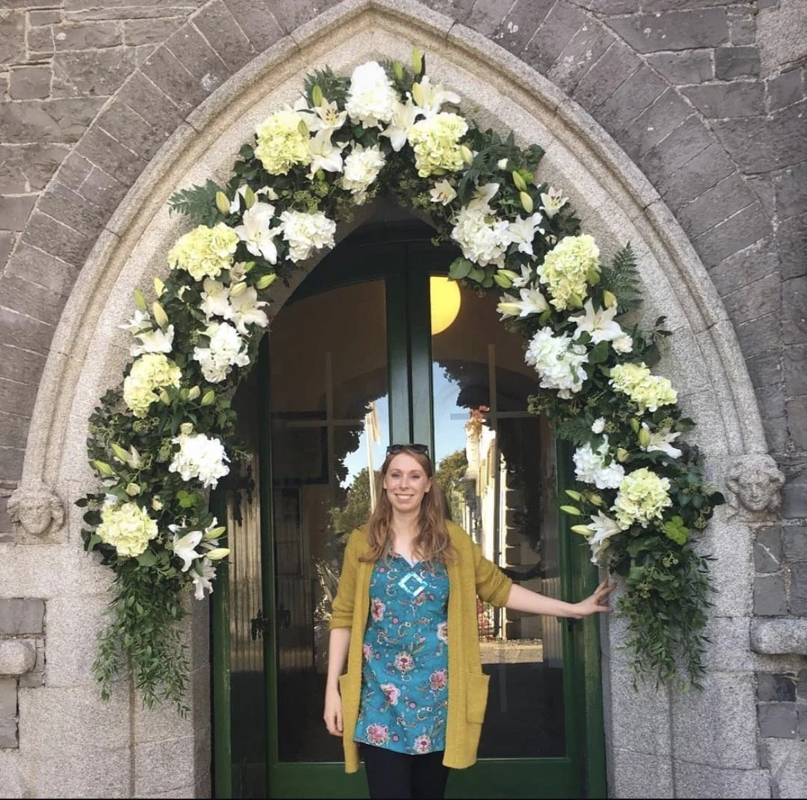 A woman standing in front of a doorway decorated with a large floral arch made of white and green flowers, touching the side of the arch with her right hand, smiling, with reflections of buildings and trees visible in the glass door.