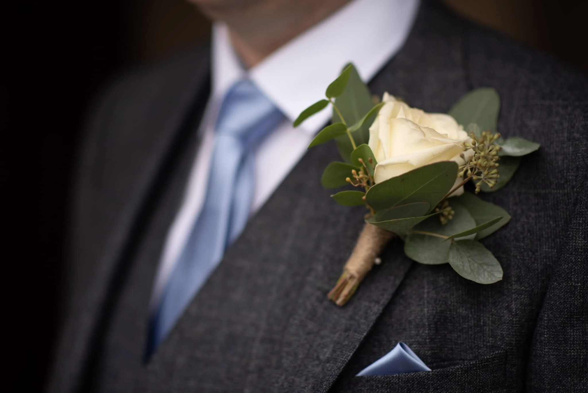 Close-up of a person's dark suit with a white rose boutonniere and greenery pinned to the lapel, wearing a light blue striped tie and pocket square.