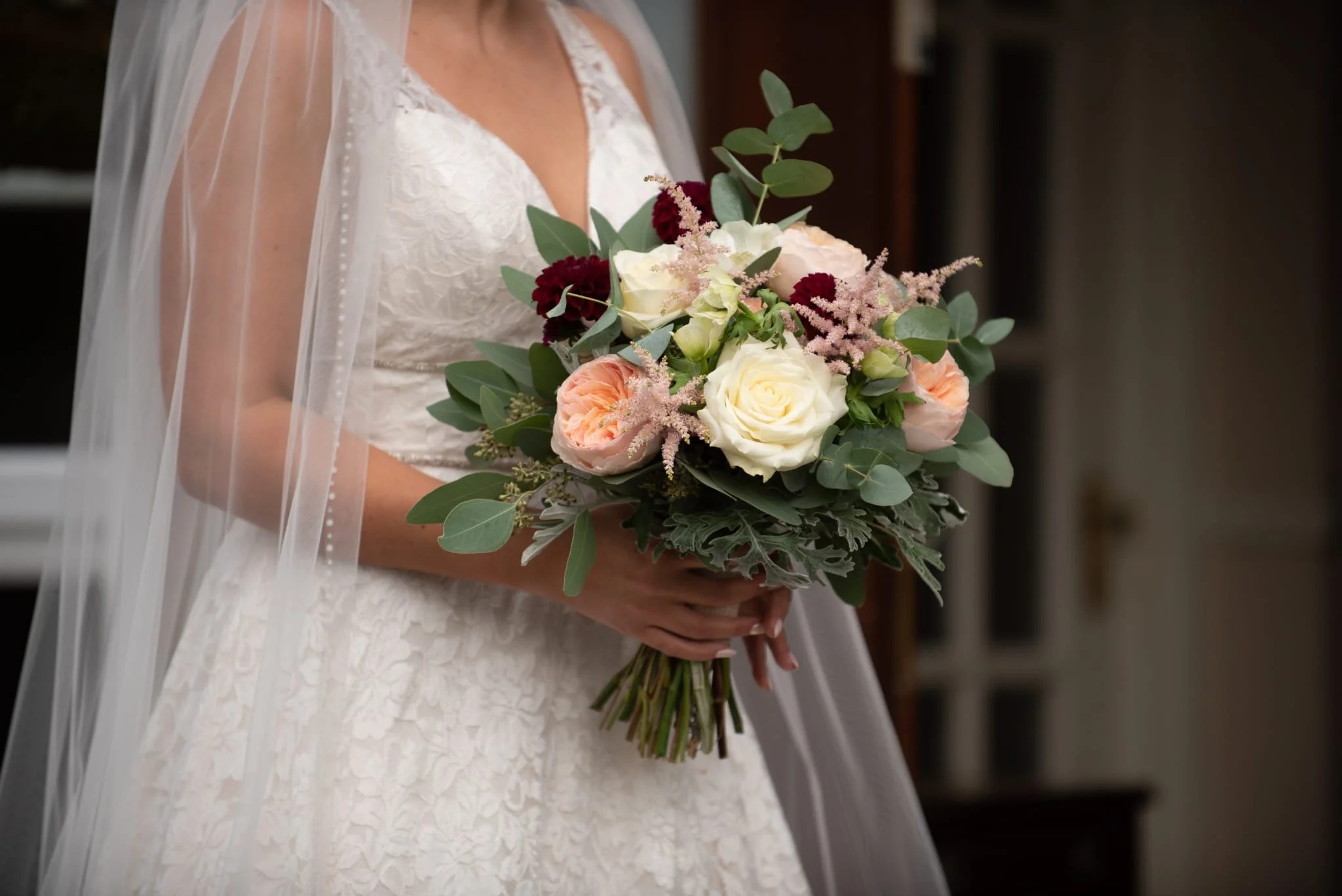 Bride wearing a lace wedding dress and veil holding a bouquet of white, pink, and burgundy flowers with green leaves.