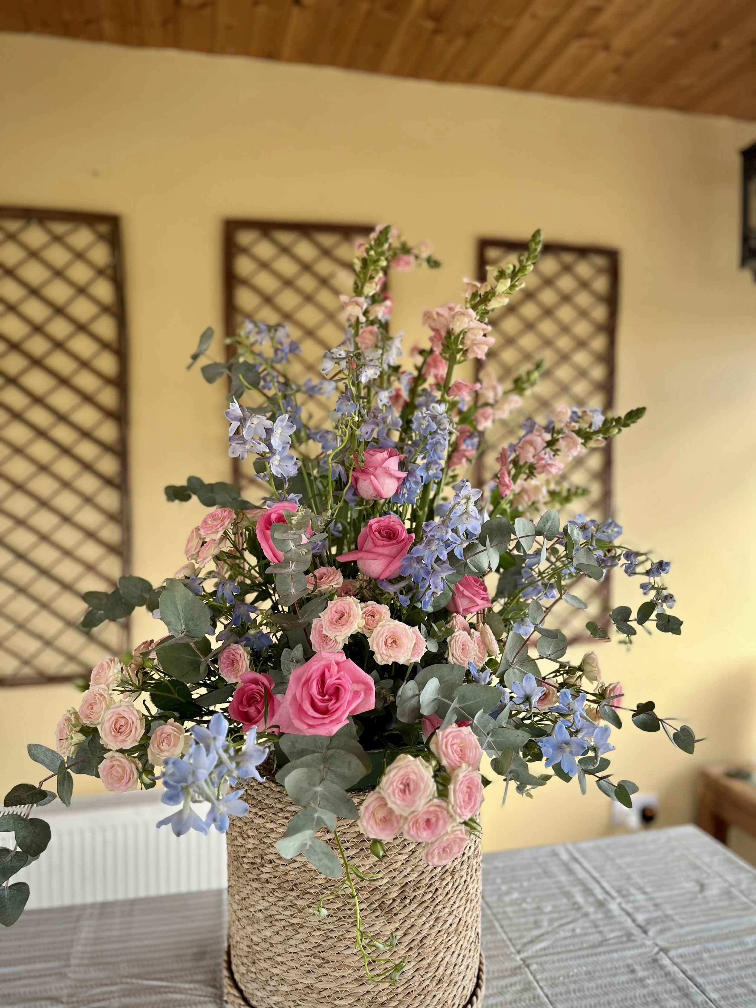 A tall floral arrangement in a woven basket contains pink roses, blue delphiniums, and light pink stock flowers, on a table with a striped tablecloth. In the background, there is a yellow wall with decorative wooden lattice pieces.
