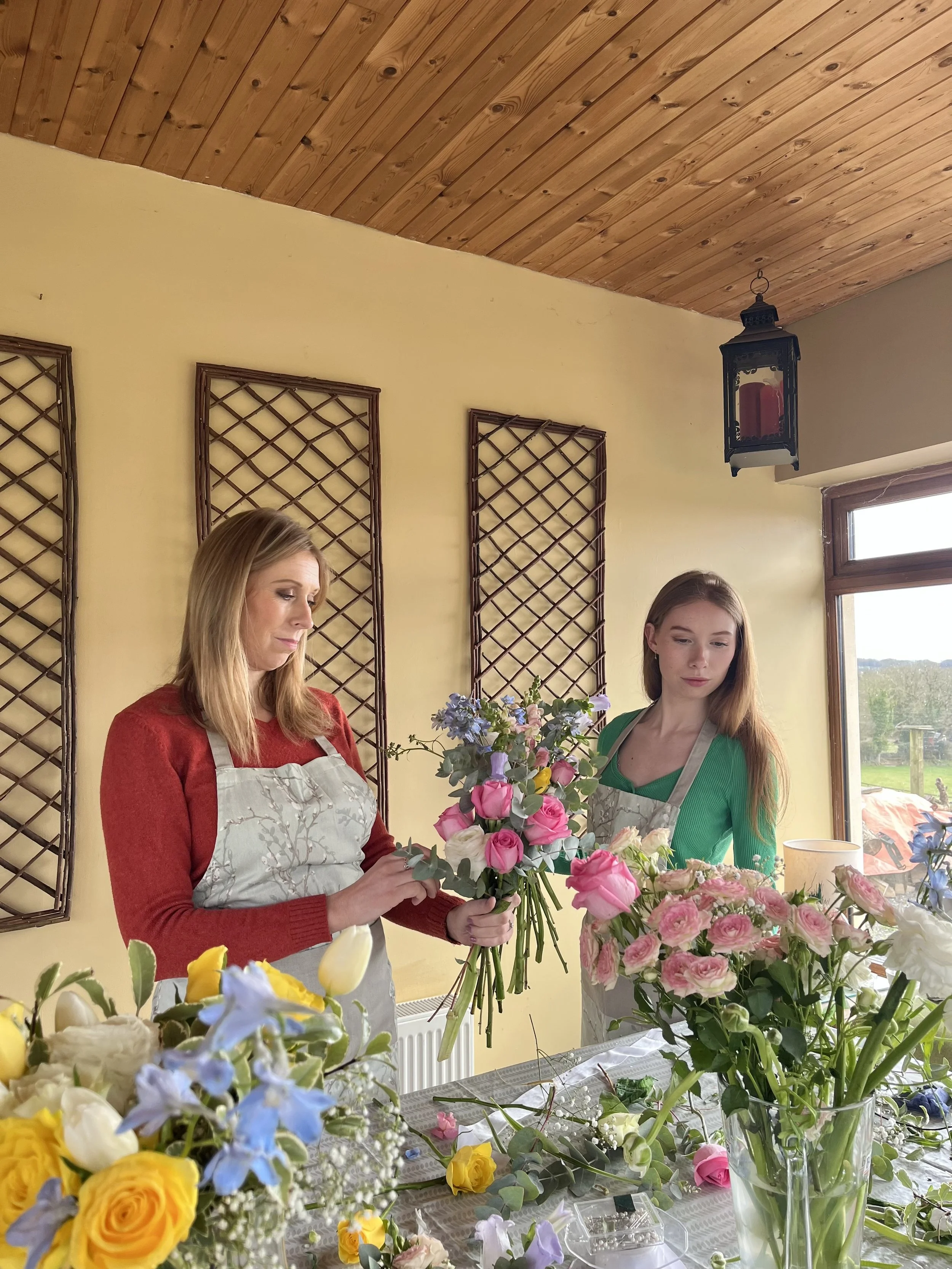 Two women arranging flowers in a sunlit room with wooden ceiling and yellow walls, surrounded by multiple vases of colorful flowers on a table.