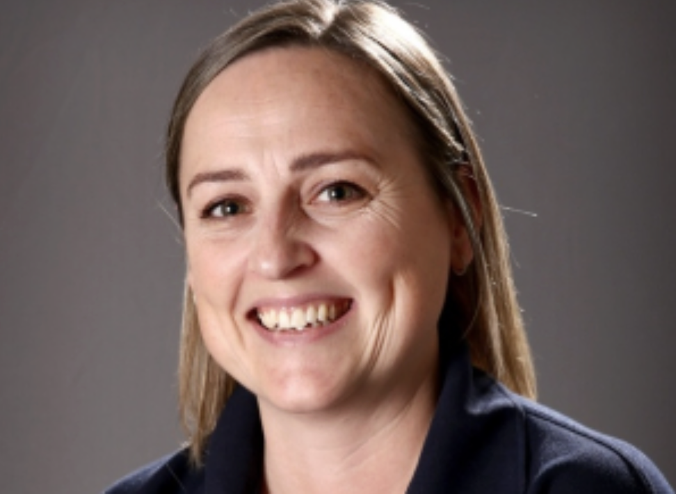 A woman with shoulder-length light brown hair smiling, wearing a dark blazer against a gray background.
