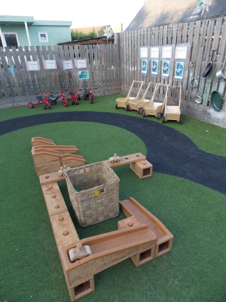 Children's outdoor play area with toy cars, tricycles, and a wicker basket, enclosed by a wooden fence with pictures and posters.