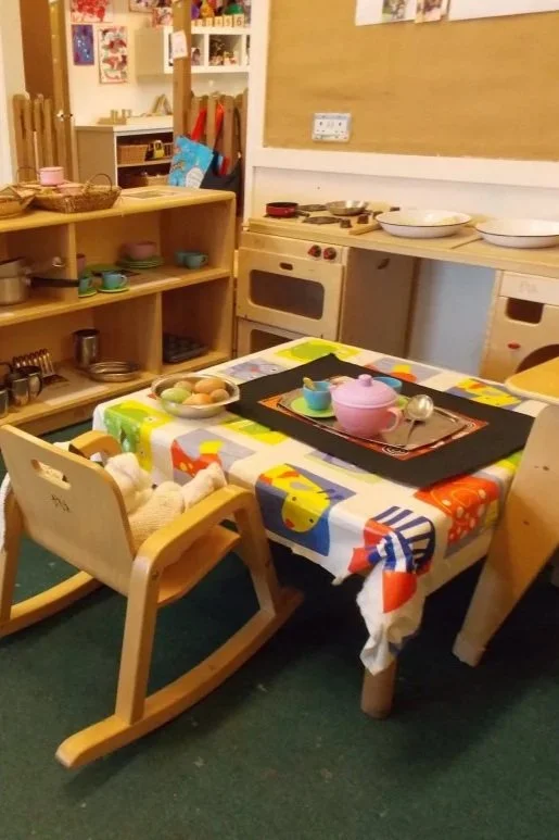 A children's play kitchen with a small wooden table covered with a colorful tablecloth, set with toy dishes and utensils, and a wooden high chair with a teddy bear seated in it. In the background, there are shelves with toy pots, pans, and dishes, and a pretend oven and stove.