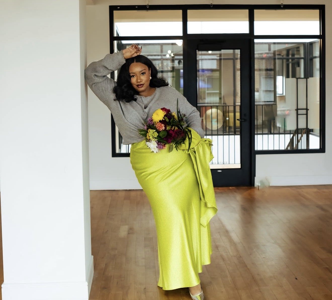 Woman with dark hair holding a colorful bouquet of flowers, wearing a lime green satin skirt and gray sweater, standing in a room with wooden floors and glass doors.