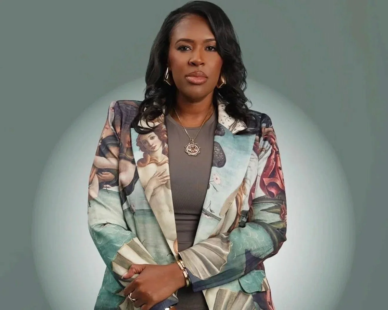 Courtney Scott with medium skin tone, black wavy hair, wearing a colorful graphic blazer with artwork and a gray top, standing with arms crossed against a plain background with a soft circular light behind her.