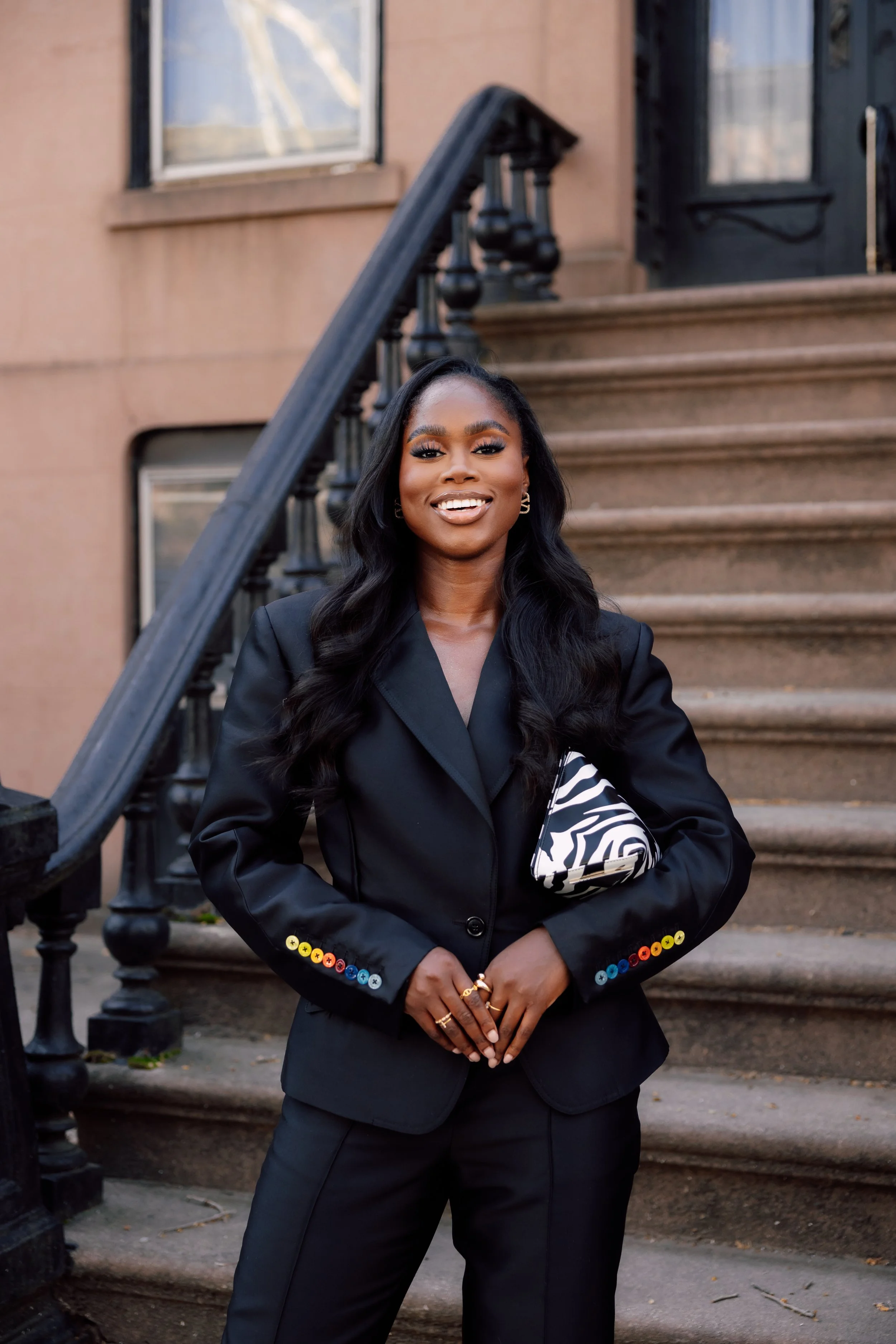 Eni Popoola in a black suit standing outdoors on a brownstone staircase, smiling, holding a black and white zebra-print clutch, wearing rings and earrings, with her long wavy black hair down.