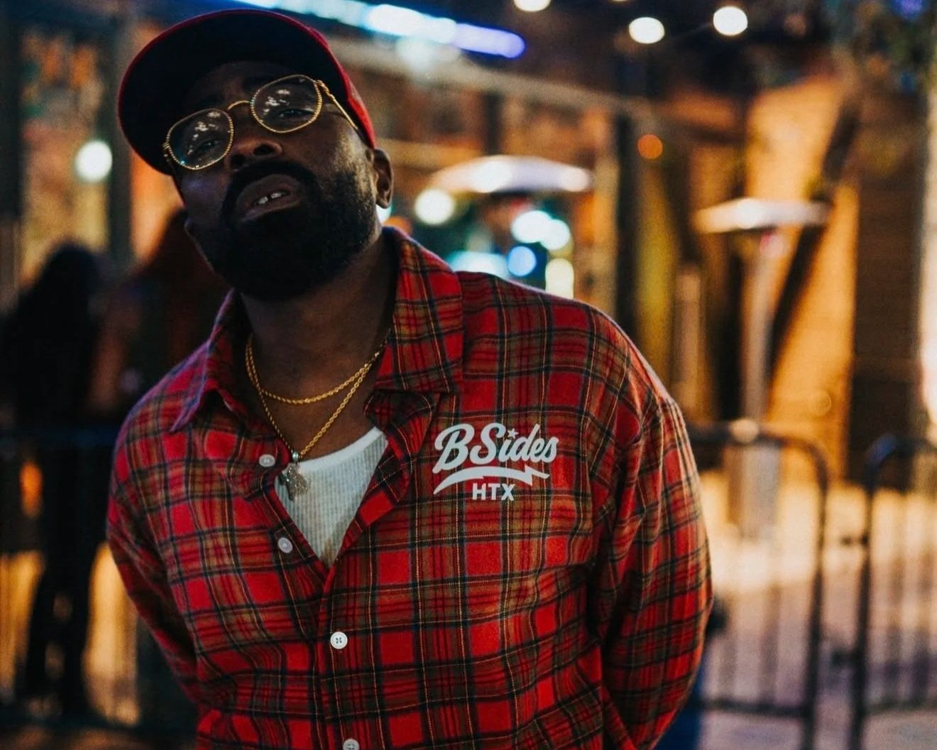 A man wearing a red baseball cap, round glasses, and gold chains, standing outdoors at night, wearing a red plaid shirt with a white patch that reads 'B Sides HTX'.