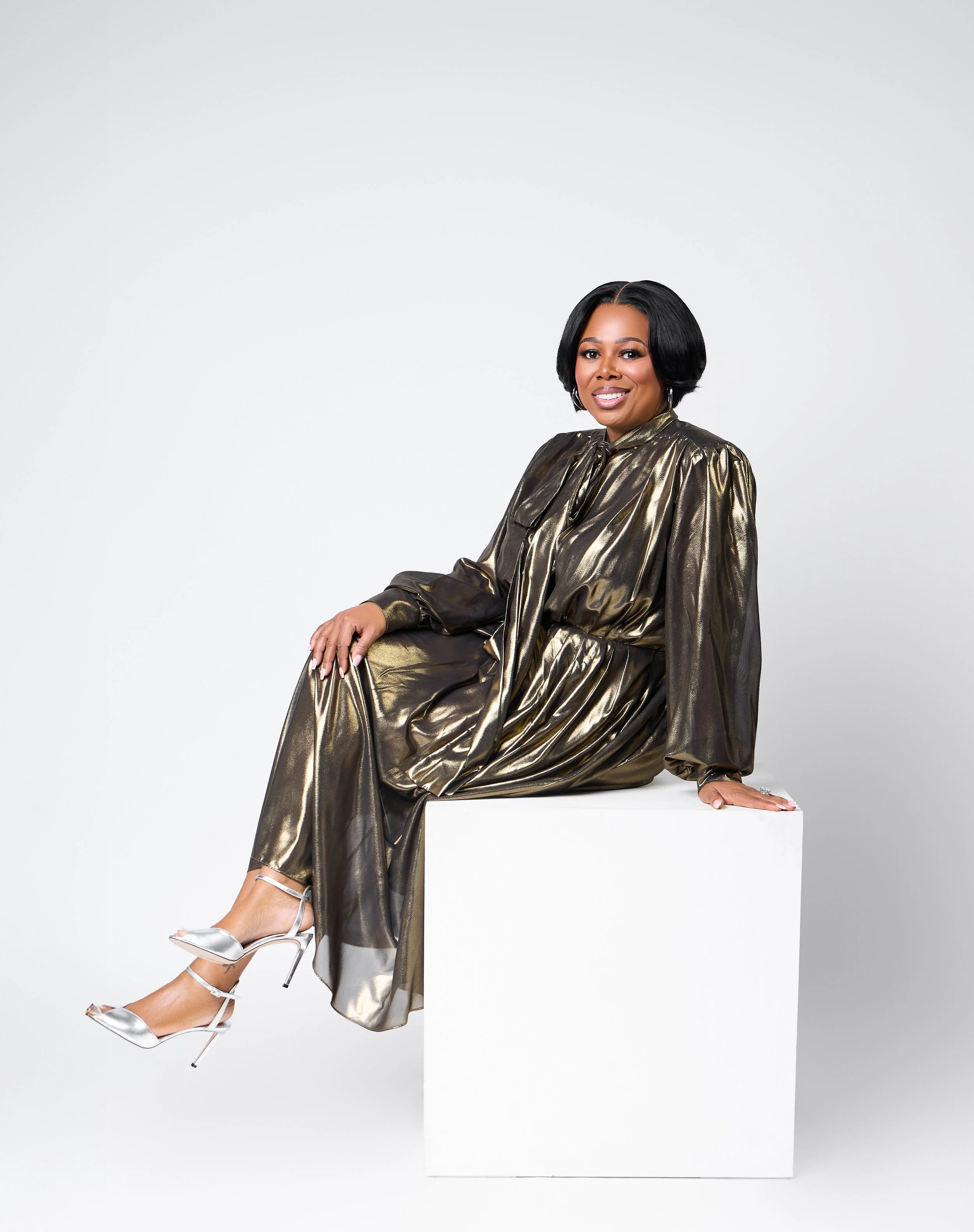 Dr. Cosha Joseph wearing a metallic gold dress and silver high heels, sitting on a white pedestal against a plain white background, smiling at the camera.