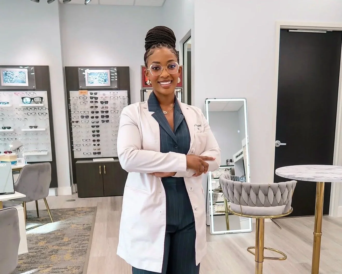 Dr. Nishan Pressley standing inside an eyewear store, smiling, wearing a white coat over dark pinstripe clothing, with displays of eyeglasses behind her and a mirror nearby.