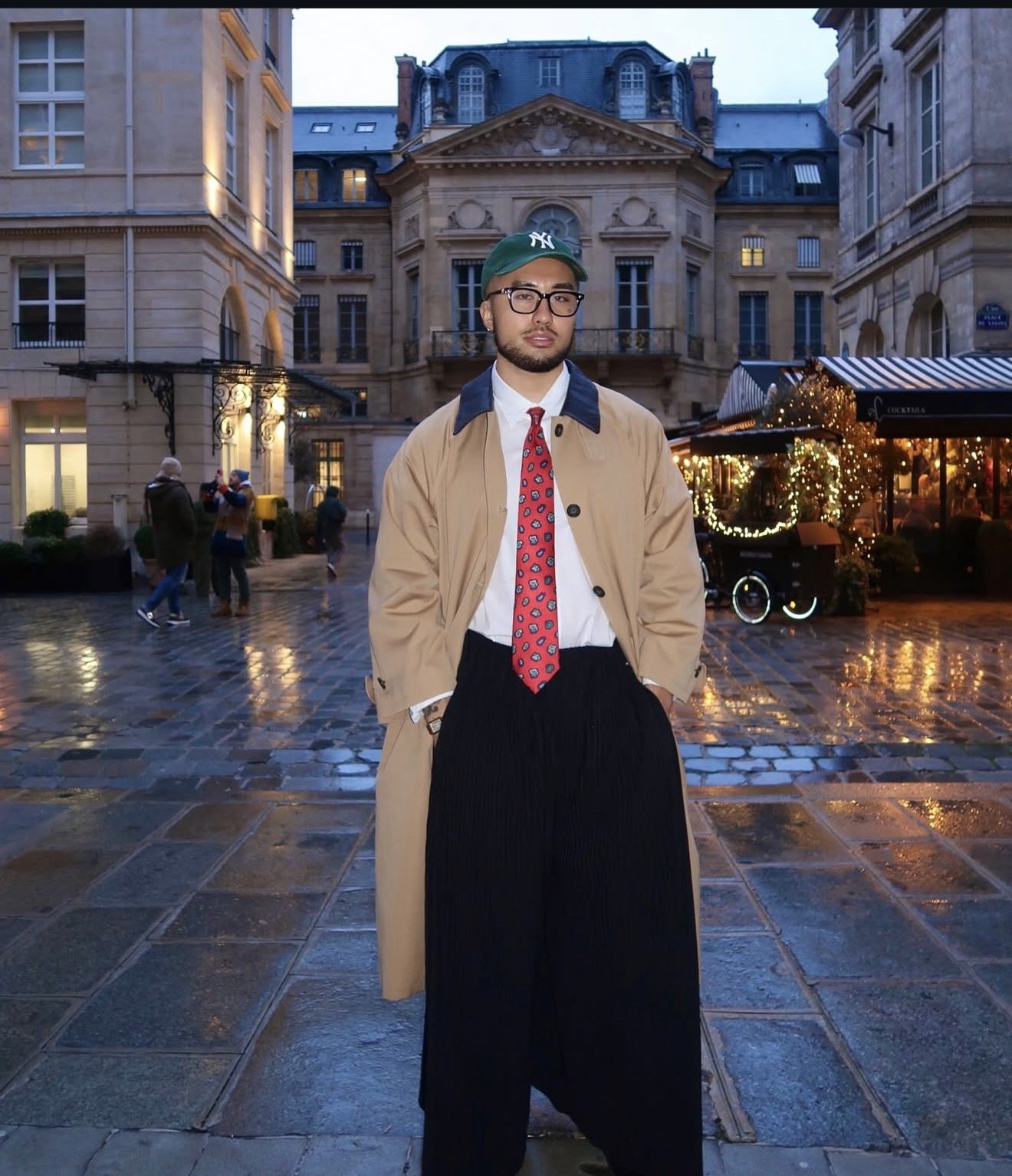 Alex Nguyen in front of historic European-style buildings during evening, wearing glasses, a beige trench coat, a white shirt, a red tie, a green cap, and black wide-leg pants, with some people and festive lights in the background.