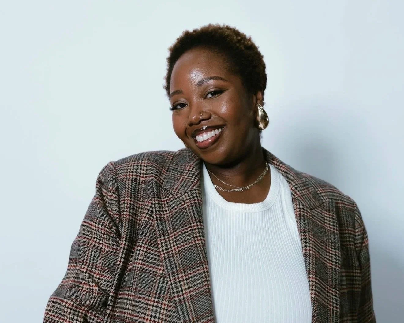 A woman with short curly hair smiling and wearing a plaid blazer, white top, and silver jewelry, against a light gray background.