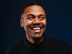 Boo Milton smiling man with short black hair, wearing a black shirt, against a dark background.