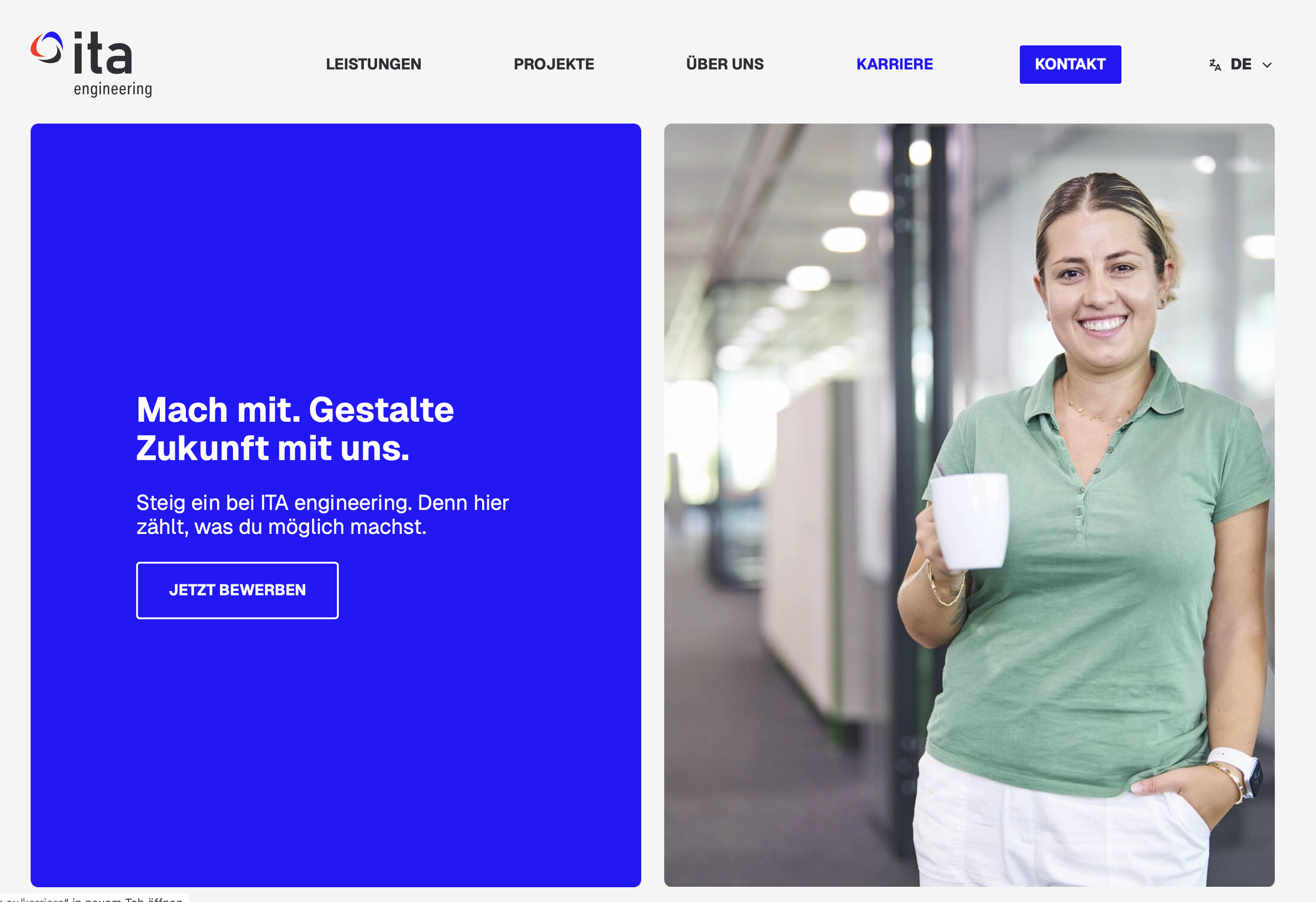 Frau mit Kurzhaarschnitt und grünem Polo Shirt, die in einem modernen Gebäude mit großen Fensterfronten einen weißen Kaffeebecher hält.