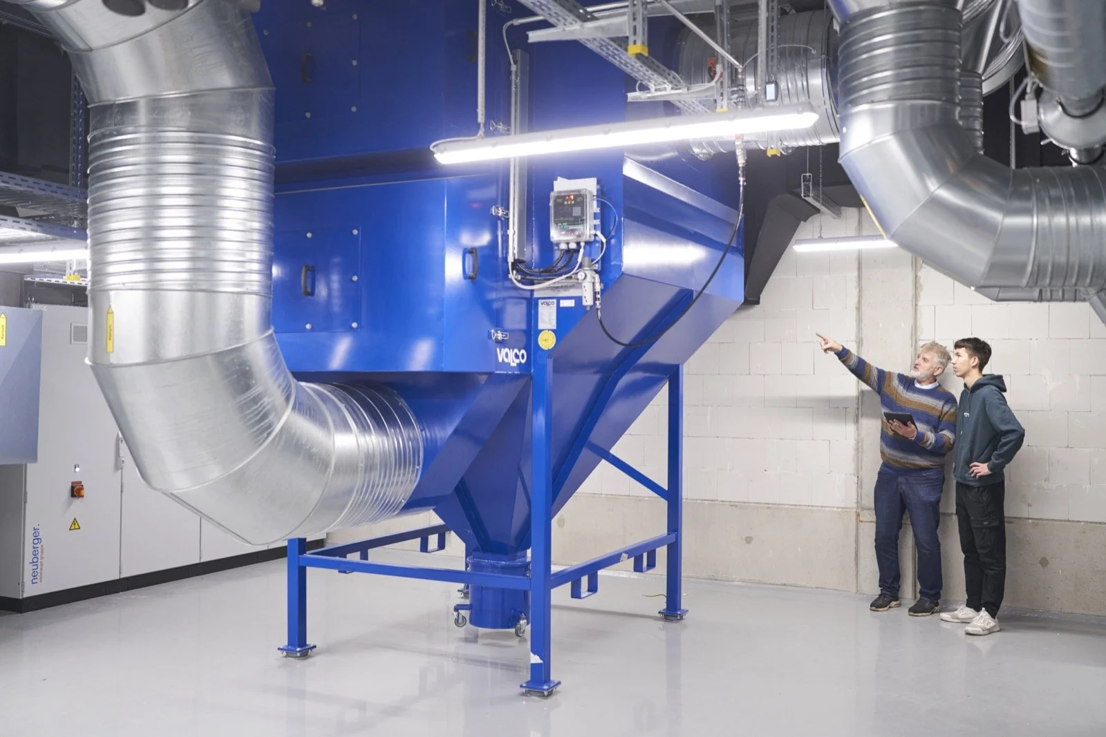 Zwei Männer, einer älter, einer jünger, stehen neben einem großen blauen Industriemaschinenstück mit metallischen Rohren, das vermutlich für die Lüftungs- oder Klimaanlagen in einer Fabrik oder Werkstatt verwendet wird, und diskutieren.
