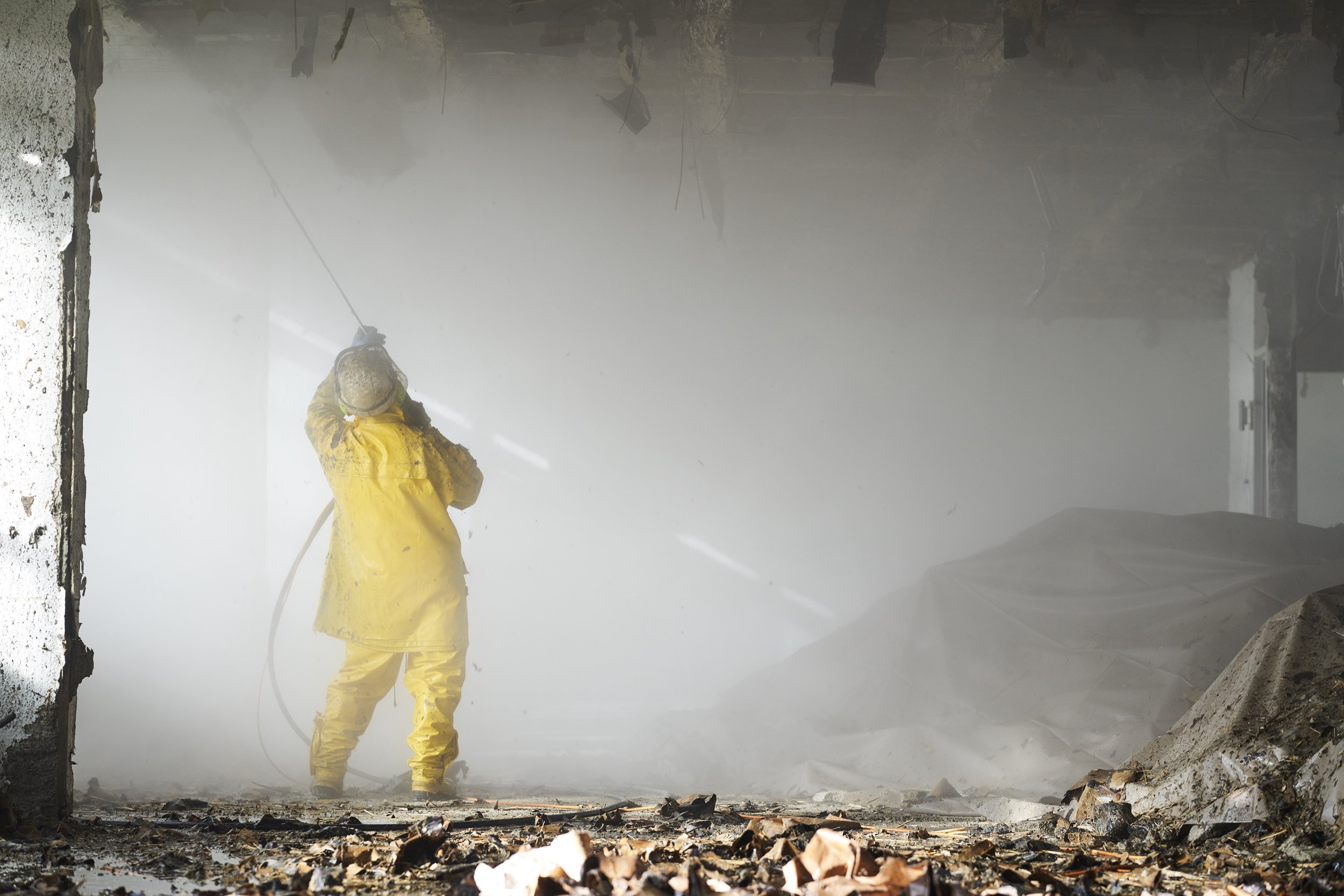 Eine Person in gelber Schutzkleidung dampft einen Raum mit einem Hochdruckreiniger, im Raum Staub und Schmutz, während die Umgebung beschädigt aussieht.  Abriss-Dokumentation der Firma Ettengruber GmbH.