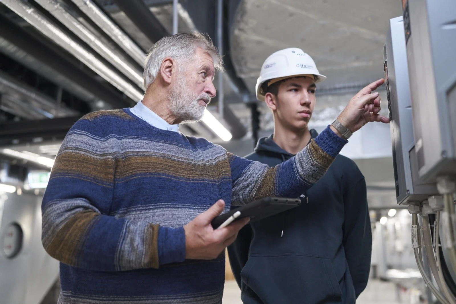 Zwei Männer, einer älter mit grauem Haar und Bart, der andere jünger mit dunklem Haar und Schutzhelm, arbeiten zusammen an elektronischen Steuerungen in einer industriellen Umgebung.