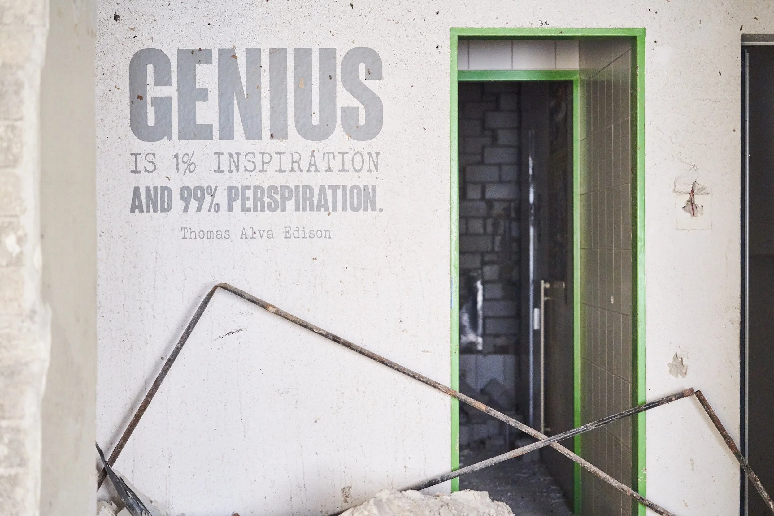 Ein Wandbild mit dem Zitat 'Genius is 1% Inspiration and 99% Perspiration' von Thomas Alva Edison, neben einer halb fertiggestellten Türöffnung in einem Gebäude im Bau, mit sichtbaren Mauern, Kabeln und Baustoffen. Abrissarbeiten der Firma Ettengrube