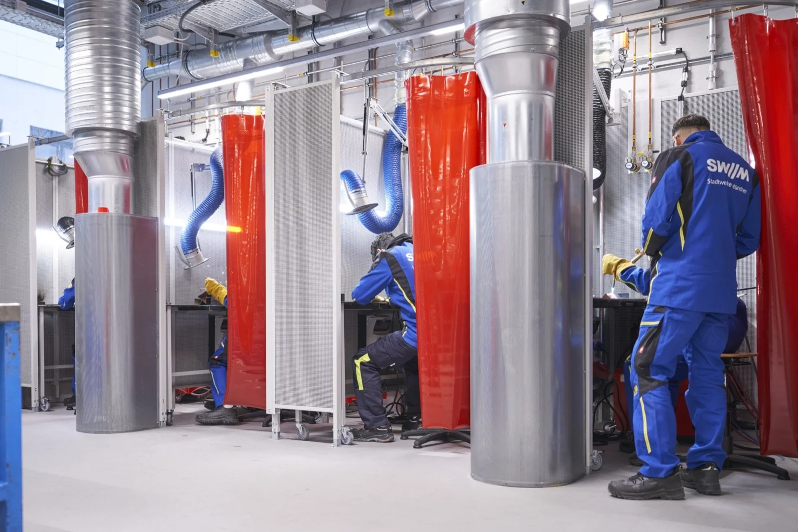 Techniker arbeiten in einem chemischen Labor mit großen runden Edelstahlgefäßen, roten Schutzabschirmungen und blauen Schläuchen, die für Sicherheits- oder Kontrollzwecke verwendet werden.