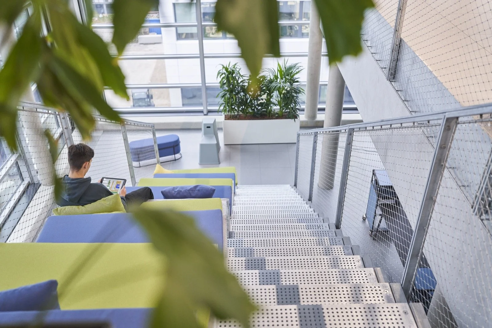 Blick auf eine moderne, helle Wartehalle oder Lounge mit Sitzbänken in Blau und Grün, einem großen Pflanzenkübel mit grünen Pflanzen, Treppe mit Metallgeländer, ein junger Mann sitzt mit einem Tablet, während im Hintergrund große Fenster mit Sicht au