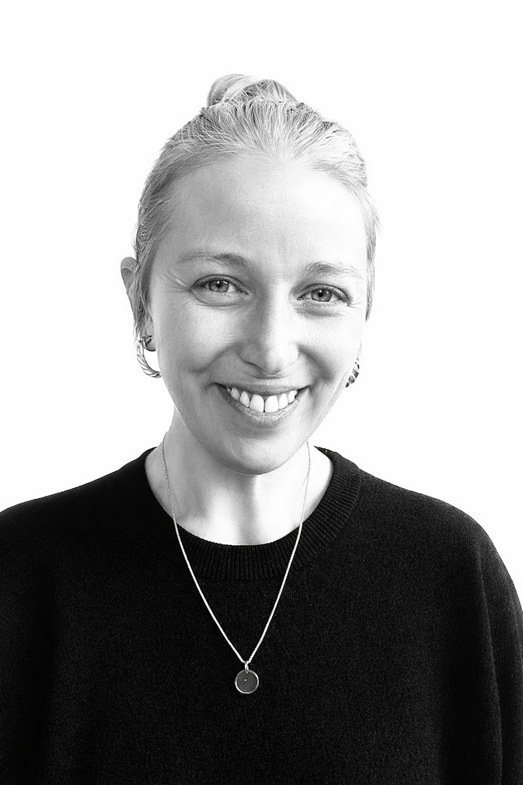 Black and white portrait of a smiling woman with short blonde hair, wearing a dark sweater and a necklace with a round pendant.