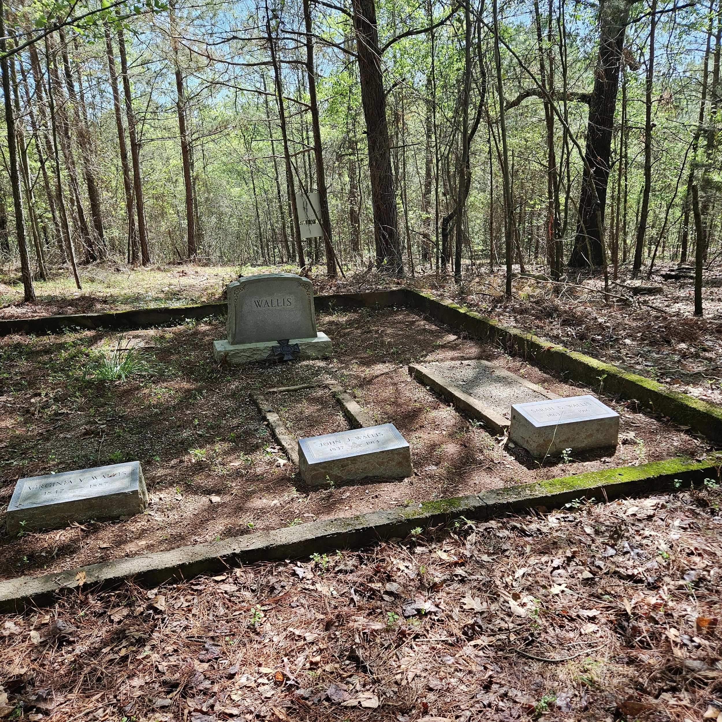 Grave site in a wooded area with four headstones. The larger headstone reads 'Wallis'. The other three are smaller and have inscriptions including names and dates, in a chain feature with a black cross decoration in front of the Wallis headstone.