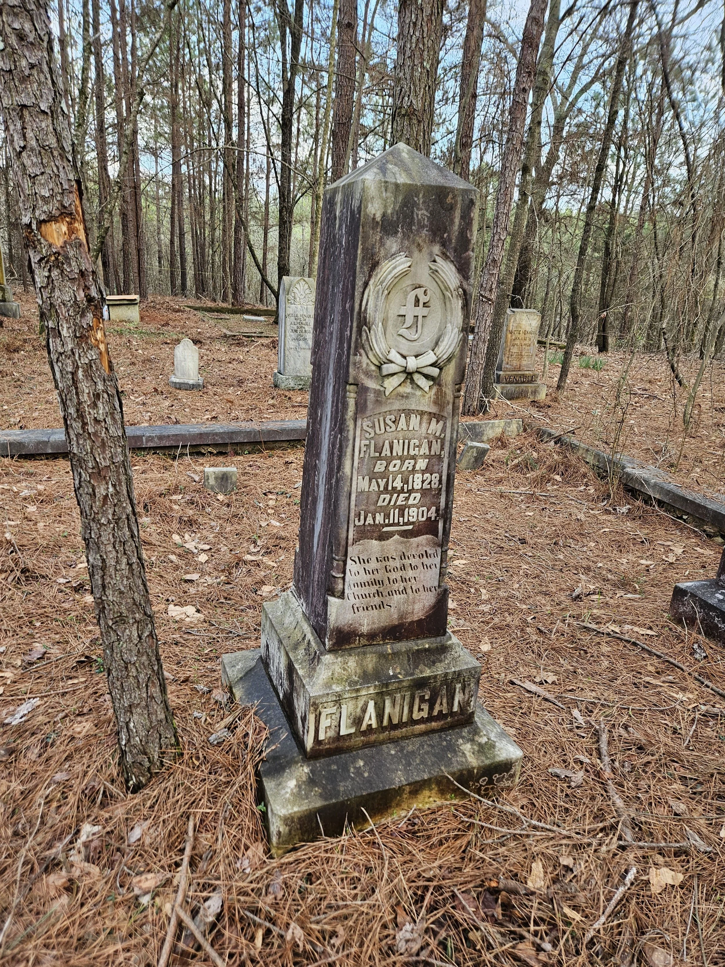 An old, moss-covered gravestone in a wooded cemetery, with other headstones in the background.