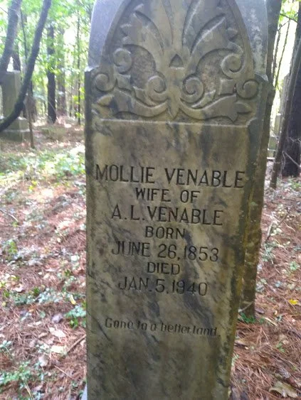 A weathered gravestone in a wooded area, inscribed with the name Mollie Venable, wife of A.L. Venable, born June 26, 1853, died January 5, 1940, with the phrase 'Gone to a better land.'