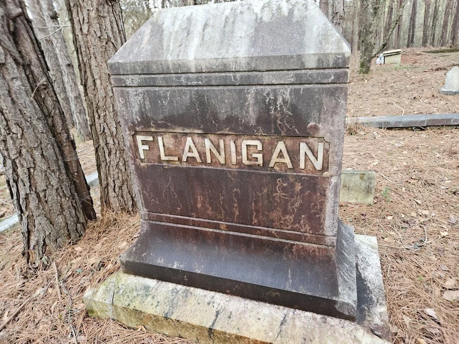 Old weathered gravestone with the name 'Flanigan' engraved in it, located in a wooded cemetery.