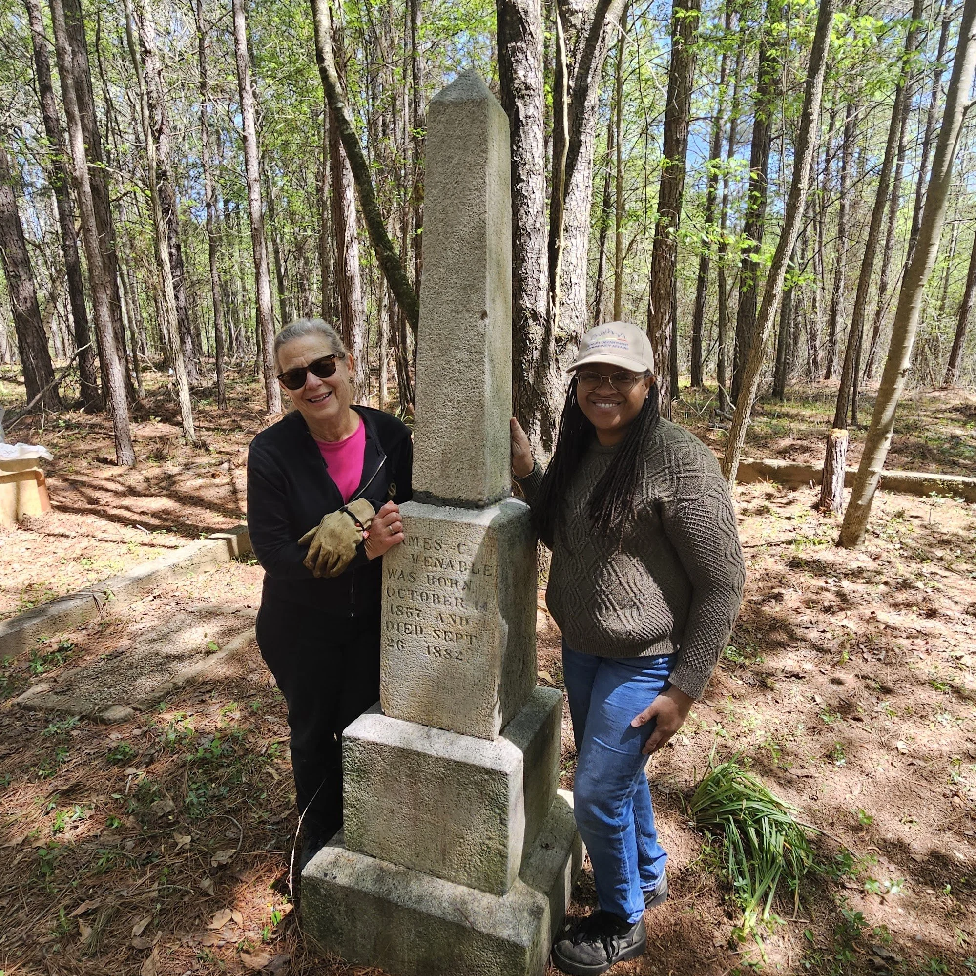 Two women smiling and standing next to a tall stone monument in a forested area.