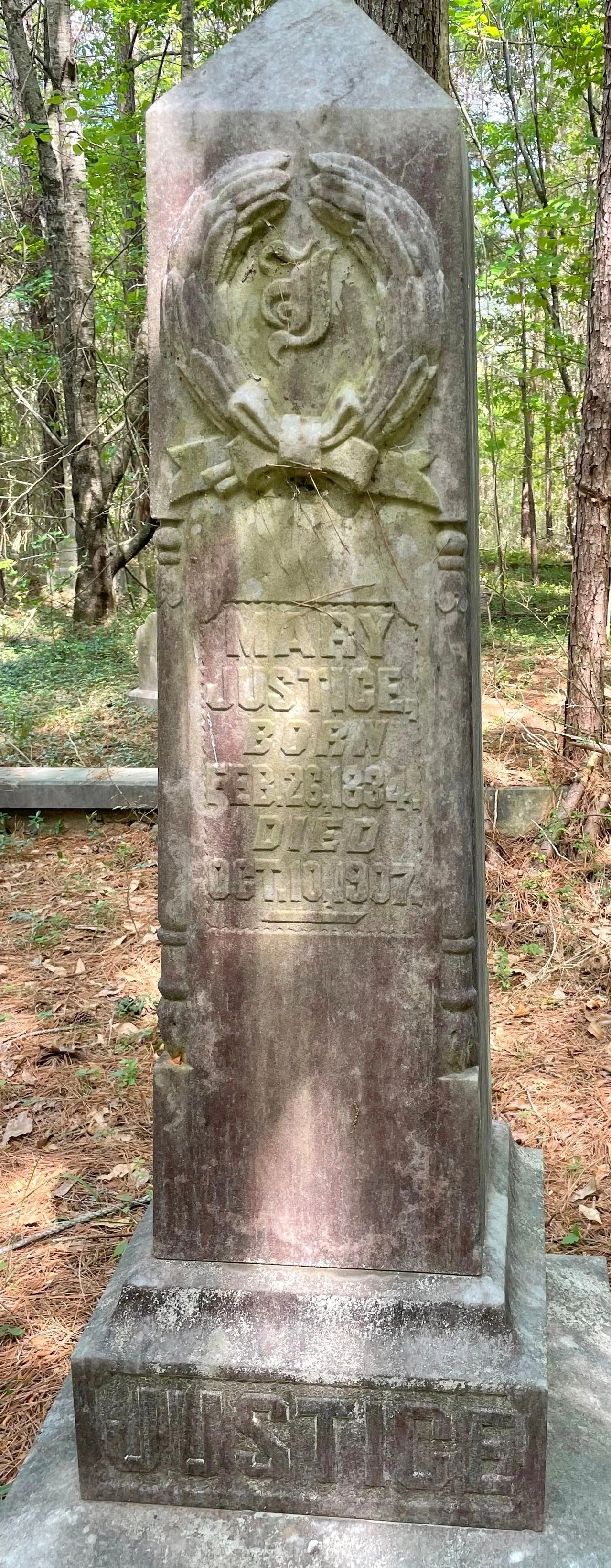 A weathered tombstone in a wooded area, inscribed with the name Justice Bowan, who died in 1904, with a carved figure of hands holding a heart at the top.