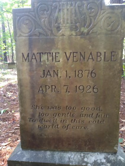A tombstone in a wooded area dedicated to Mattie Venable, who was born on January 1, 1876, and died on April 7, 1926. The tombstone has an inscribed verse: 'she was too good, too gentle and fair, to dwell in this cold world of care.'