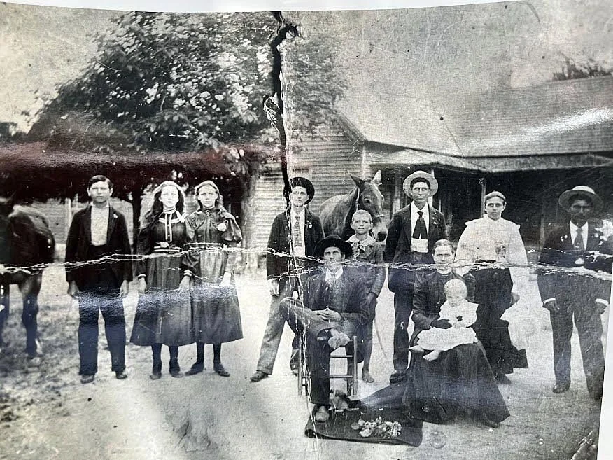 A vintage black-and-white photograph of a family gathered outdoors in front of a wooden building. The group includes men, women, young girls, and a baby, with one man sitting on a chair and a woman holding the baby. An older woman is seated on the ground, and a horse is visible in the background. The photo appears aged and slightly damaged.