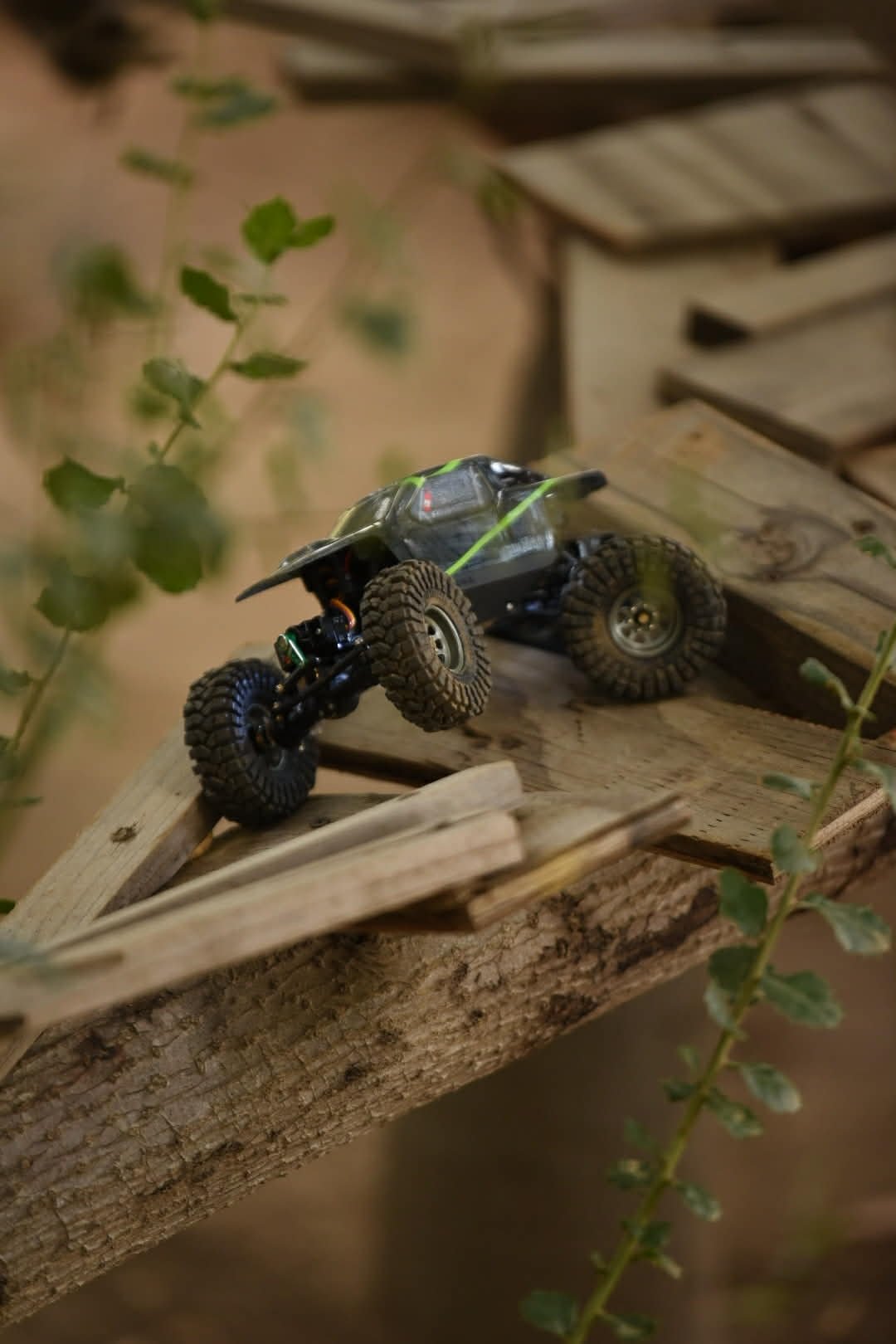 A small toy off-road vehicle with large tires and a black body, positioned on a piece of wood with plants around.