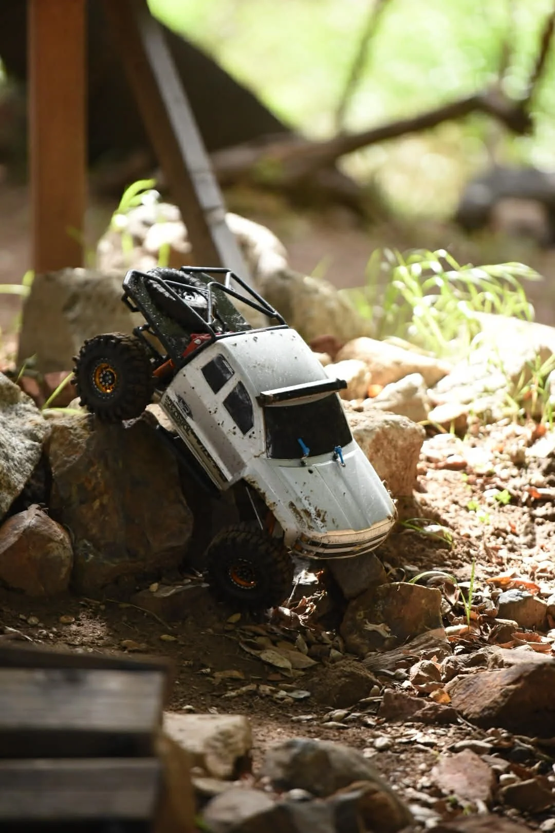 A toy RC off-road truck climbing over rocks on a dirt trail in a grassy outdoor area with sunlight filtering through trees.
