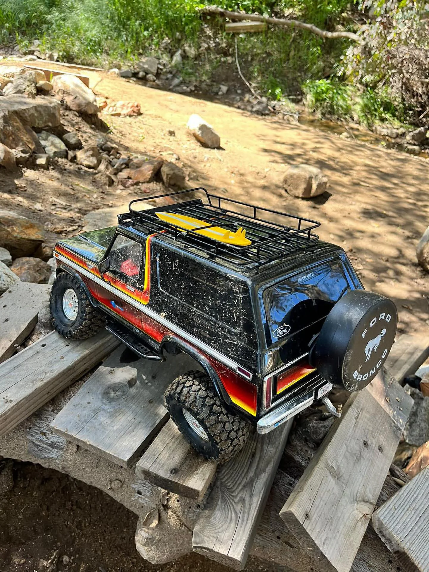 A black and red toy Ford Bronco remote control car with a yellow surfboard on its roof, placed on wooden planks outdoors near a dirt path and rocks.