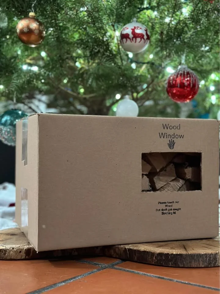A cardboard box with 'Wood Window' and a small window cutout showing wood scraps inside. The box is on a wooden surface with a decorated Christmas tree in the background.