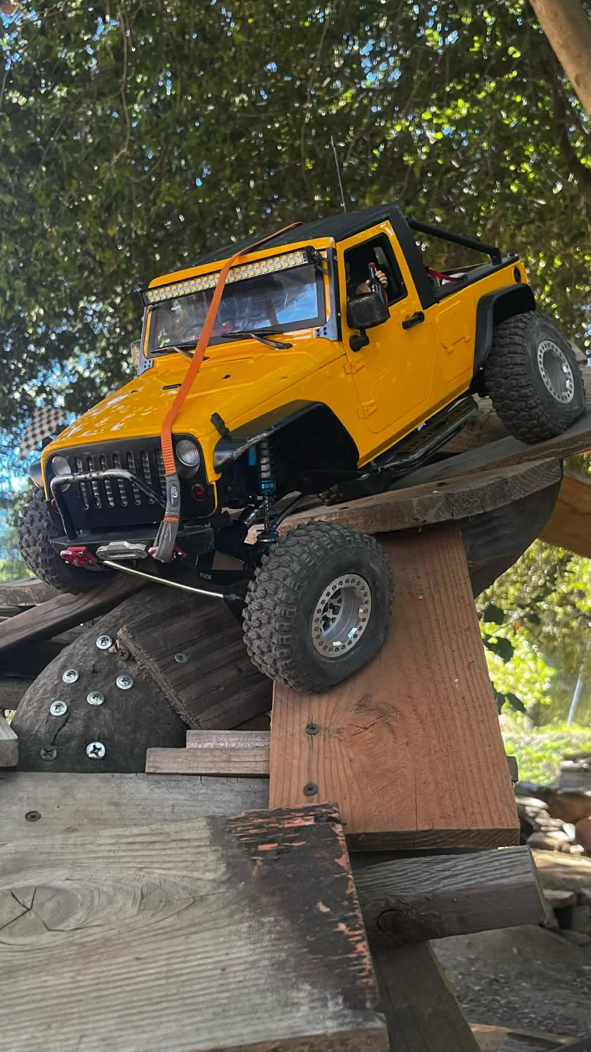 A yellow remote-controlled off-road vehicle on a wooden ramp outdoors, with trees and blue sky in the background.