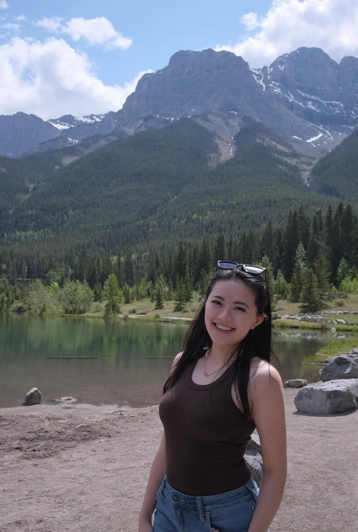 A young woman smiling at a lake with a mountain range and pine trees in the background.