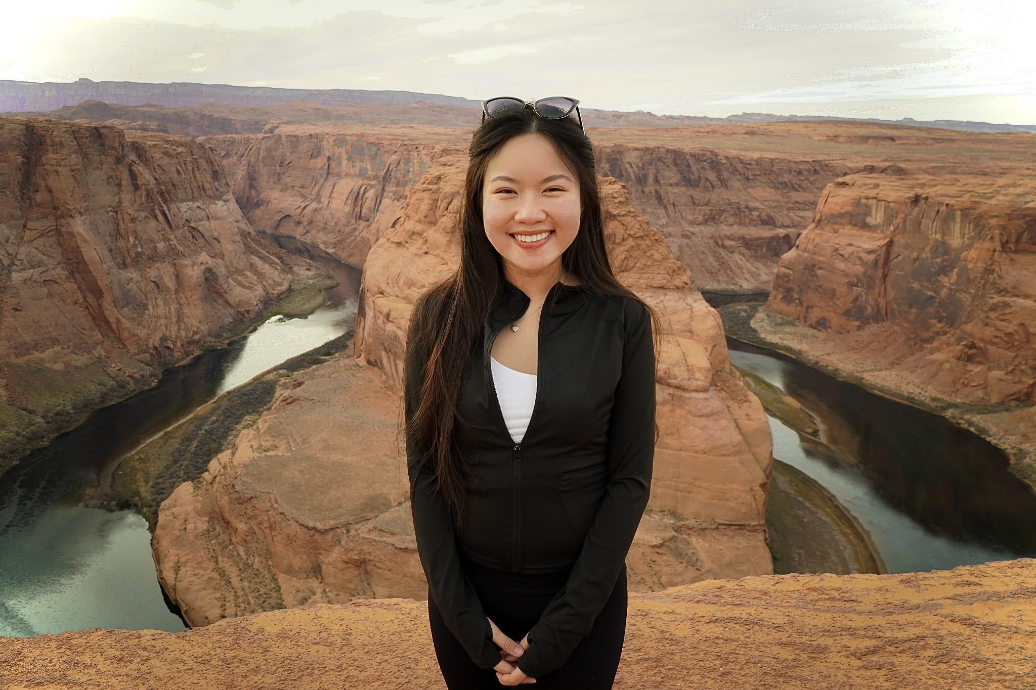 A young woman with long dark hair and sunglasses on her head smiling at the camera, standing in front of a canyon with a winding river below.
