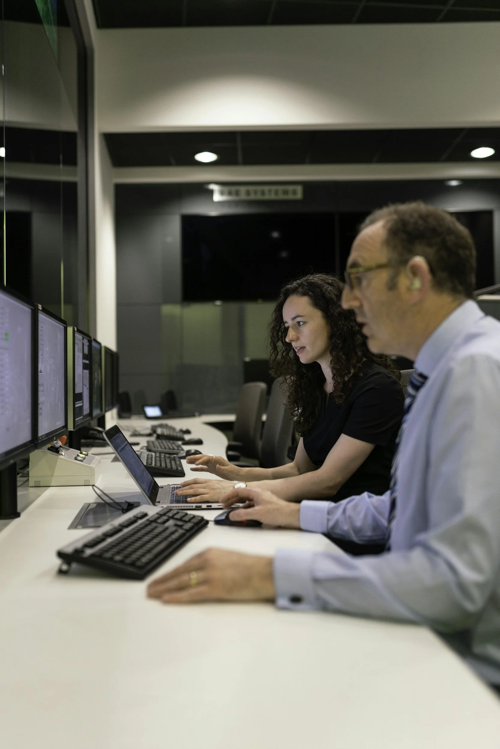 Two people working at a control room with multiple computer monitors.