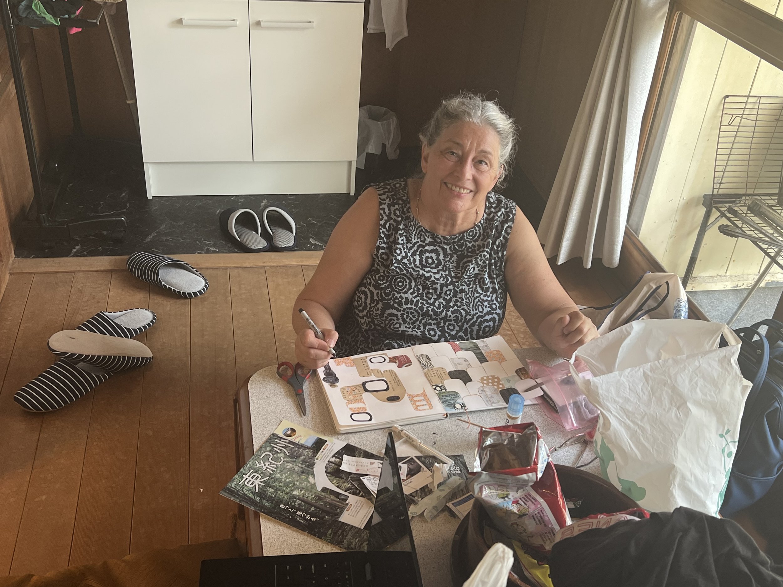 An elderly woman sitting at a table, smiling, surrounded by sewing and craft supplies, with slippers on the floor near a doorway, in a room with a wooden floor and a window with curtains.