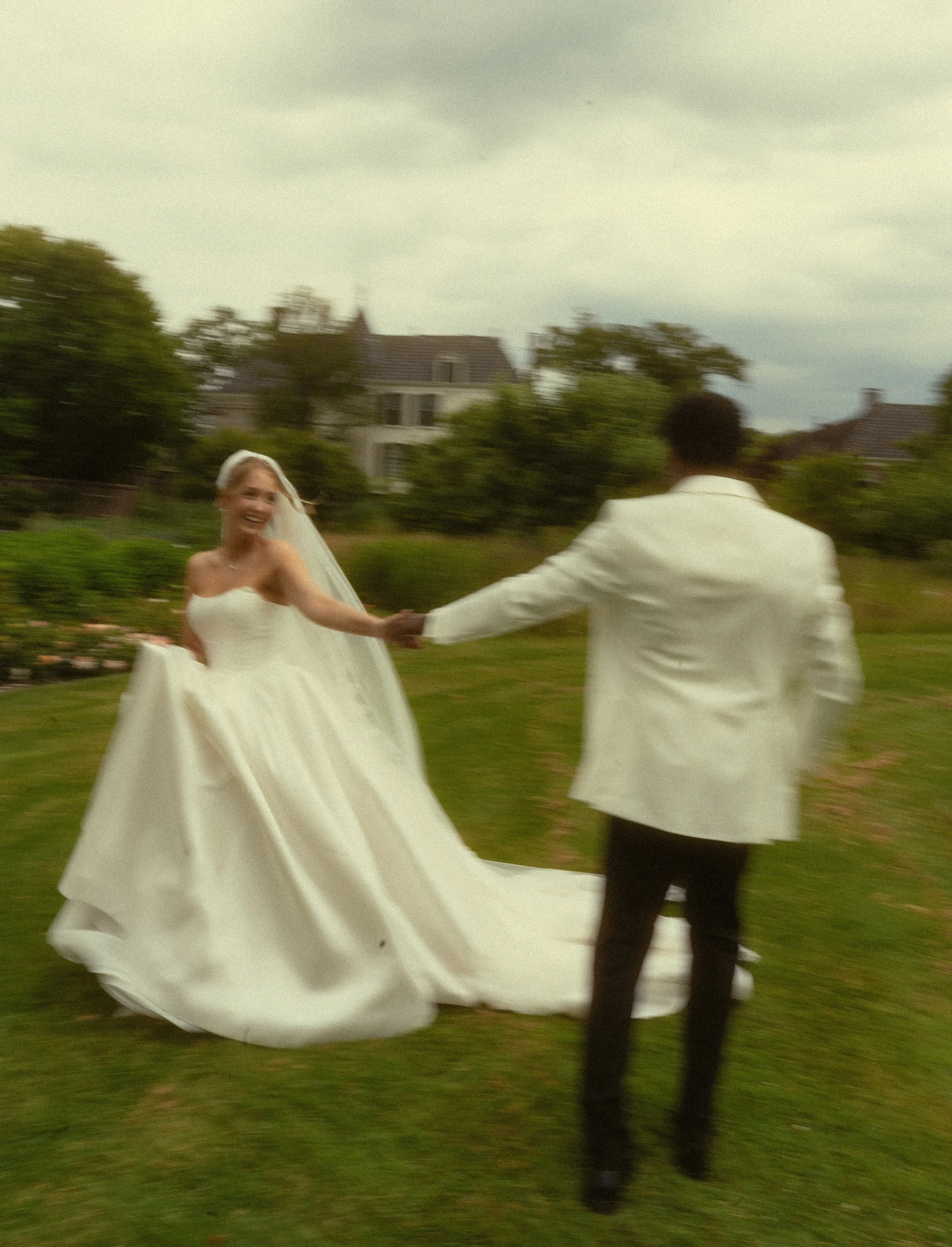 Een bruid en bruidegom dansen buiten op gras met een huis en bomen op de achtergrond.