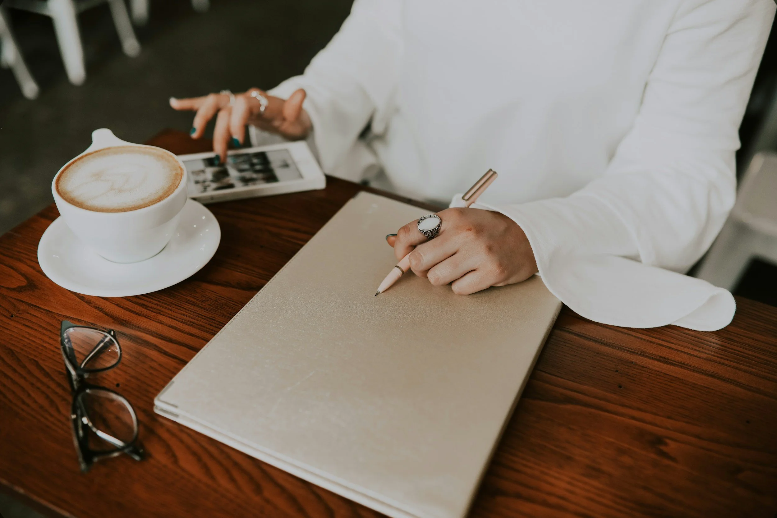 A person in a white long sleeve shirt is sitting at a wooden table, writing in a beige notebook with a white pen. On the table, there are glasses, a cup of coffee with foam art, a smartphone, and some photos.