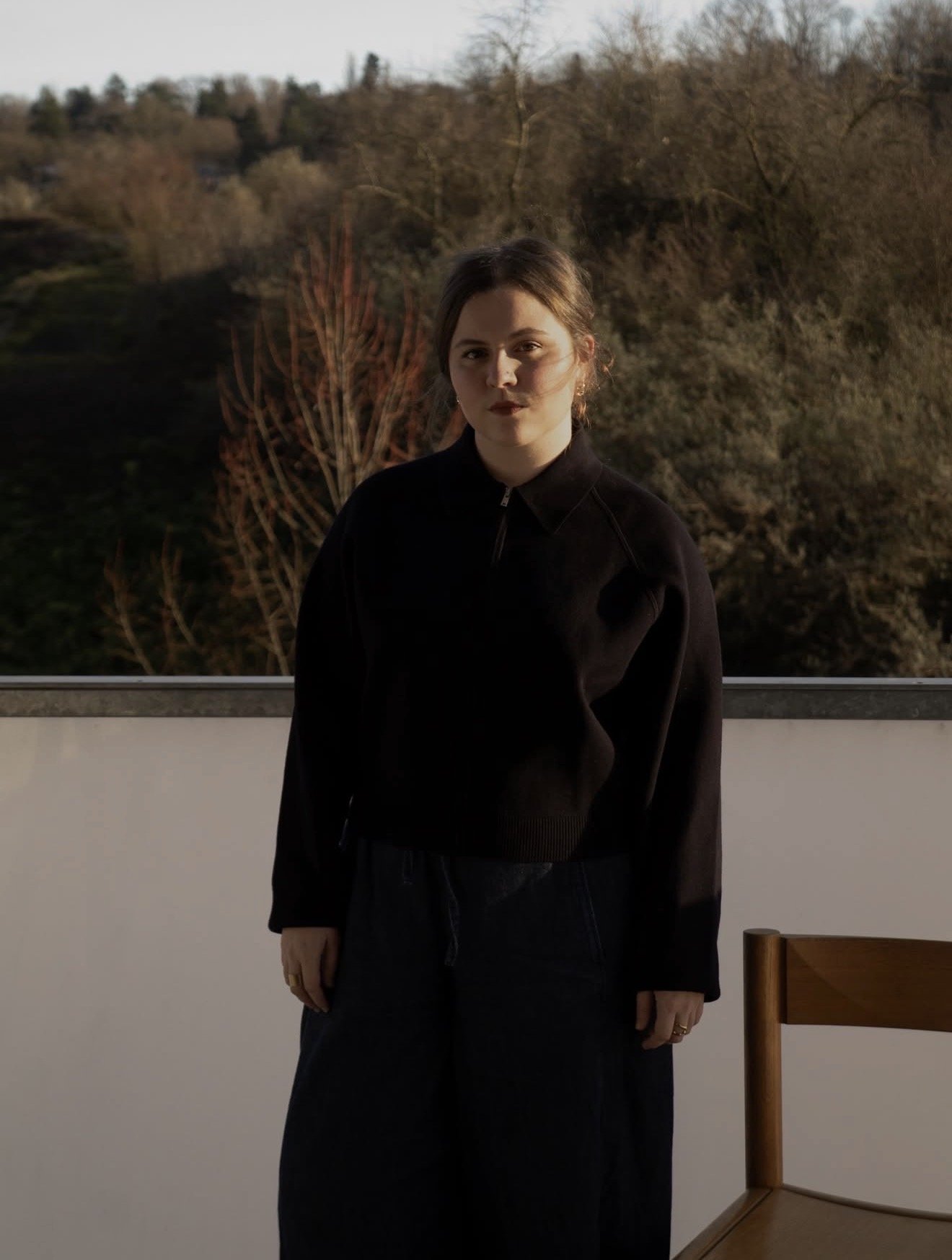 A young woman with dark hair standing indoors in front of a large window showing a landscape of trees and a hill, wearing a black zip-up jacket and dark pants.