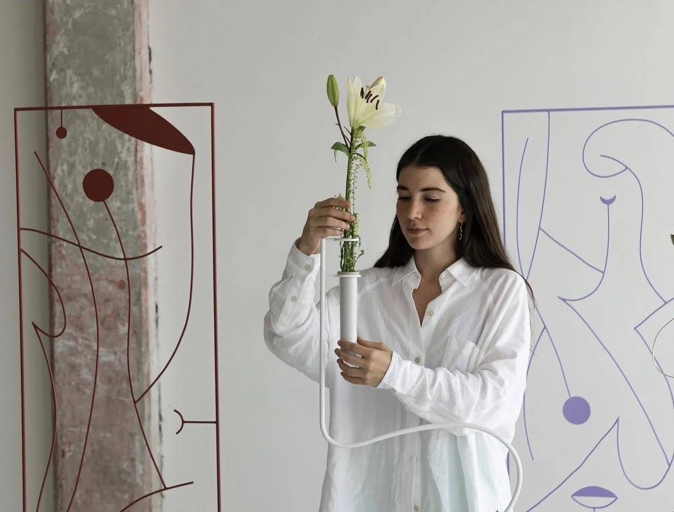 Woman in white shirt holding a tall glass vase with white lilies and greenery.