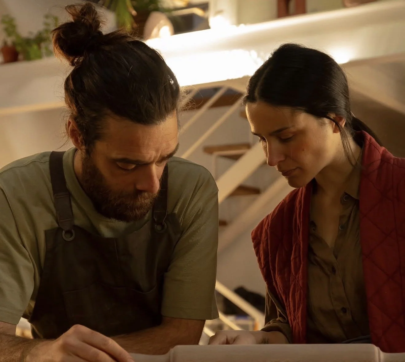 A man and woman are closely looking at something, possibly a book or paper, with a staircase and plants in the background.